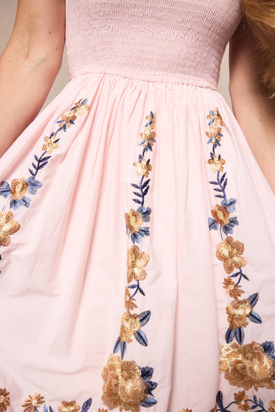 A woman wearing the Ivy City Co Hattie Dress in Embroidered Spring Floral—a light pink, maternity-friendly midi with a smocked bodice and gold and blue floral embroidery—gently holds out the sides of her dress.