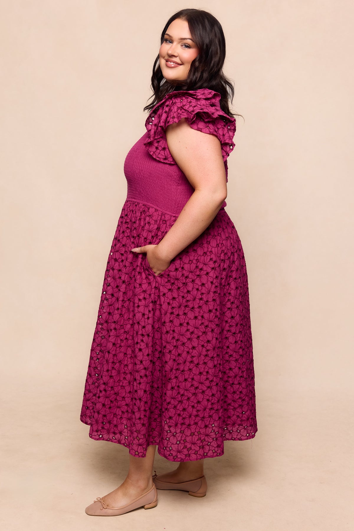A woman smiles sideways, wearing the Ivy City Co Hattie Dress in Magenta—a sleeveless, maternity-friendly, midi-length dress with ruffled shoulders and a lace skirt. She pairs it with nude flats, one hand in her pocket against a neutral background.