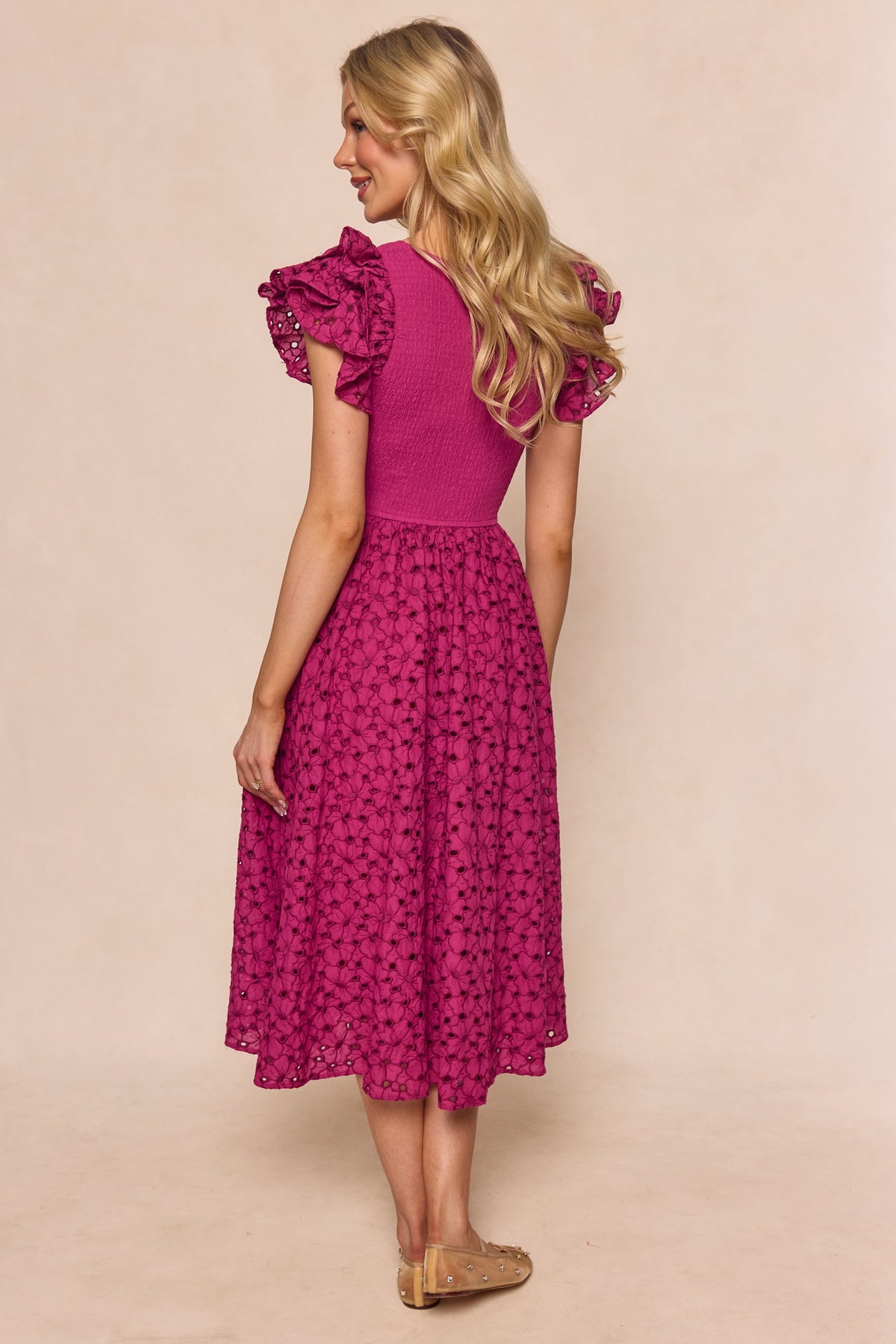 A woman with long blonde hair stands on a light background wearing the Ivy City Co Hattie Dress in Magenta, a midi-length style with short ruffled sleeves and a textured embroidered floral pattern, paired with beige flats.