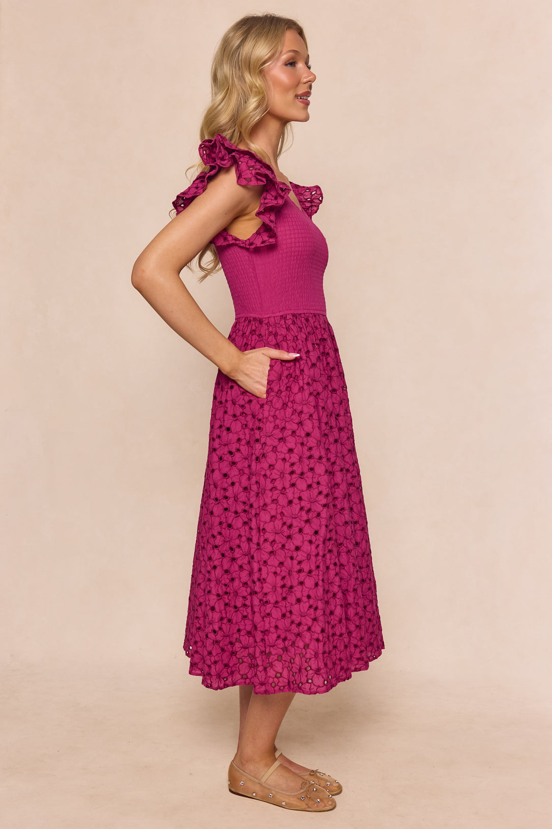 A woman with blonde wavy hair stands in profile wearing the Ivy City Co Hattie Dress in Magenta—a sleeveless, embroidered midi dress with a textured floral lace skirt and ruffled shoulders. She pairs it with nude flats and has one hand in her pocket.