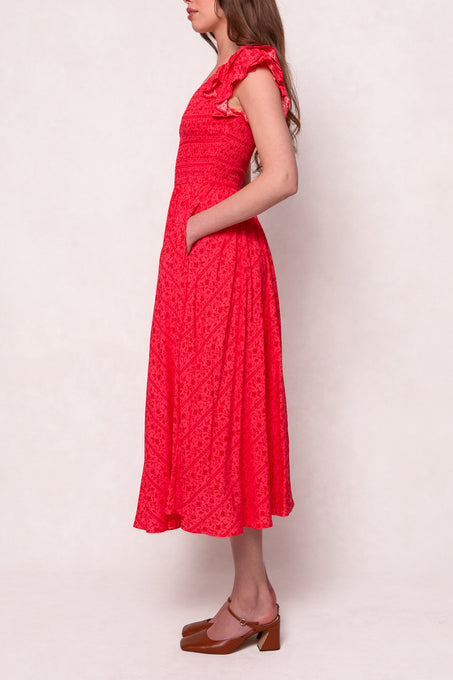 A woman stands sideways in the Ivy City Co Hattie Dress in Crimson Pink—a cotton midi dress with ruffled shoulders, short sleeves, and pockets—paired with brown low-heeled shoes against a light background.