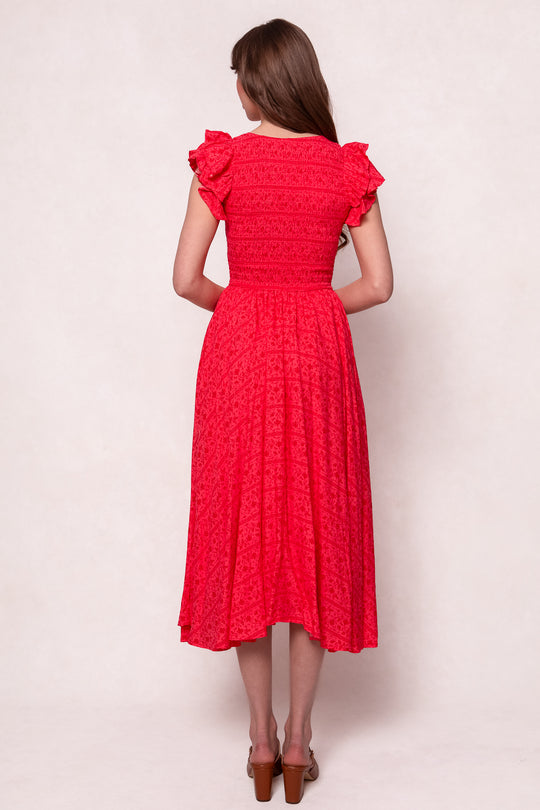 A woman with long brown hair wears the Ivy City Co Hattie Dress in Crimson Pink—a bright, knee-length cotton midi dress with ruffled sleeves and a fitted bodice—paired with brown heeled sandals, standing against a light background.