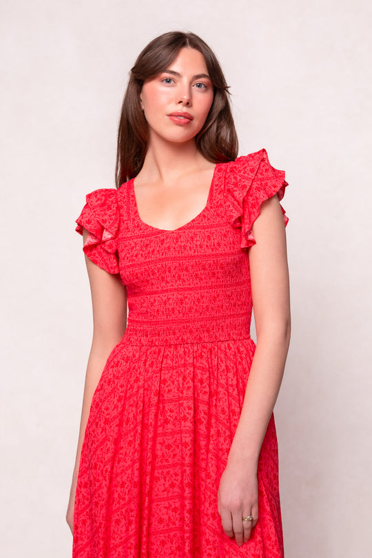 A woman with long brown hair wears the Ivy City Co Hattie Dress in Crimson Pink, a short-sleeved cotton midi with ruffled shoulders and subtle pattern, standing against a light neutral background.