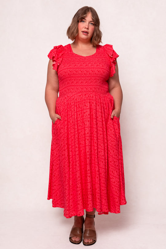 A woman with short brown hair wears the Ivy City Co Hattie Dress in Crimson Pink, an ankle-length cotton midi with ruffled sleeves and a subtle pattern. She faces forward, hands in side pockets, paired with brown sandals.