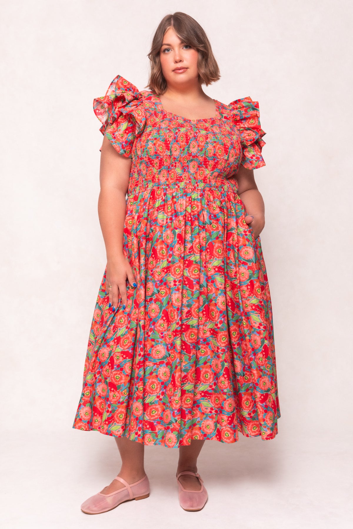 A woman in the Ivy City Co Hattie Dress in Poppy—a cotton, poppy-print, puff-sleeve midi with a smocked bodice and full skirt—stands against a light background. She has shoulder-length brown hair and wears pink flats.