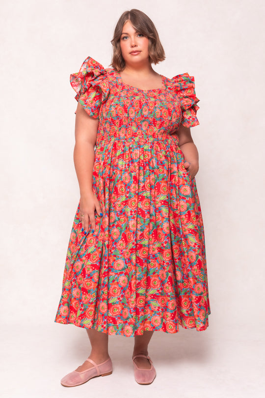 A woman in the Ivy City Co Hattie Dress in Poppy—a cotton, poppy-print, puff-sleeve midi with a smocked bodice and full skirt—stands against a light background. She has shoulder-length brown hair and wears pink flats.