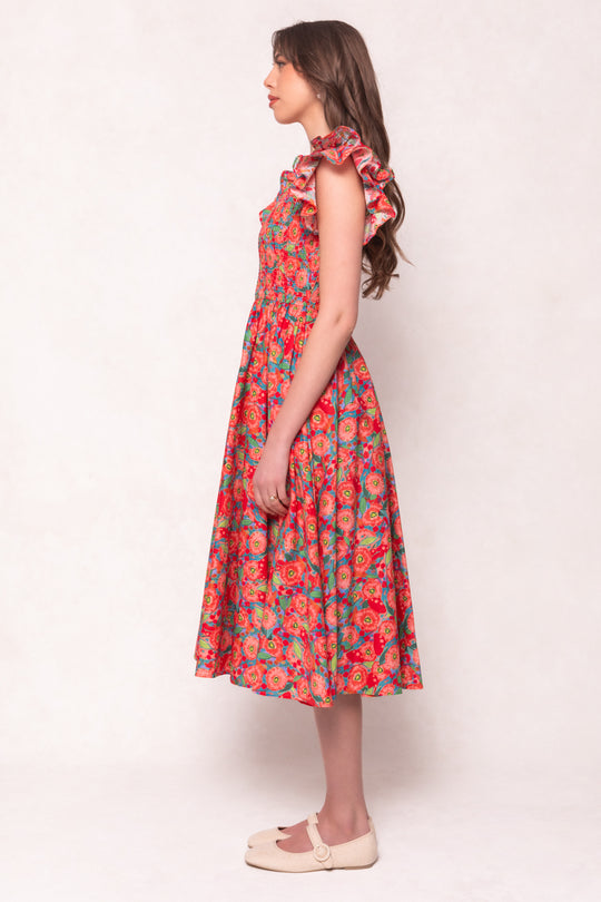 A young woman with long, wavy brown hair stands in profile against a light background, wearing the Ivy City Co Hattie Dress in Poppy—a cotton midi dress with ruffled shoulders—paired with light flat shoes.