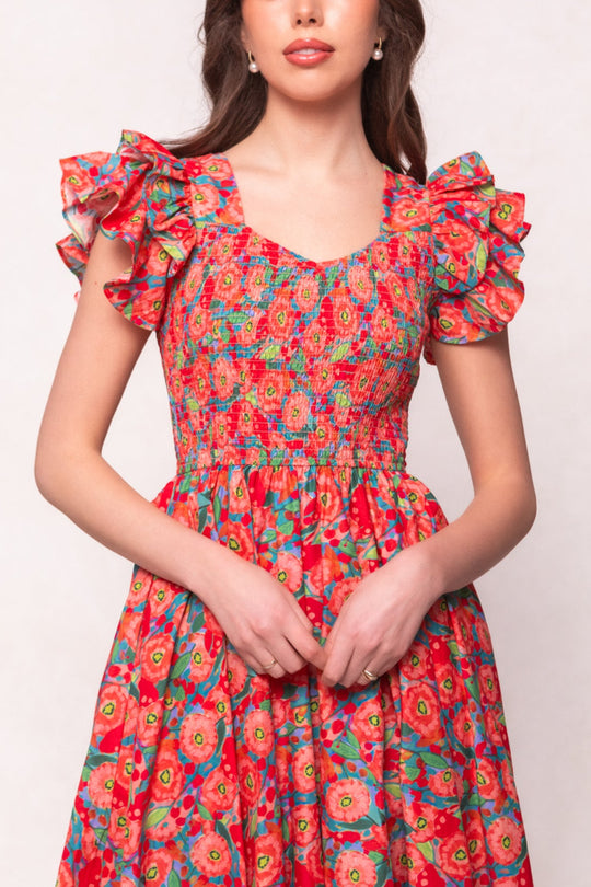A woman wearing the Ivy City Co Hattie Dress in Poppy—a cotton midi dress with a vibrant red poppy print, smocked bodice, and ruffled sleeves—stands against a light background with her hands gently clasped in front.