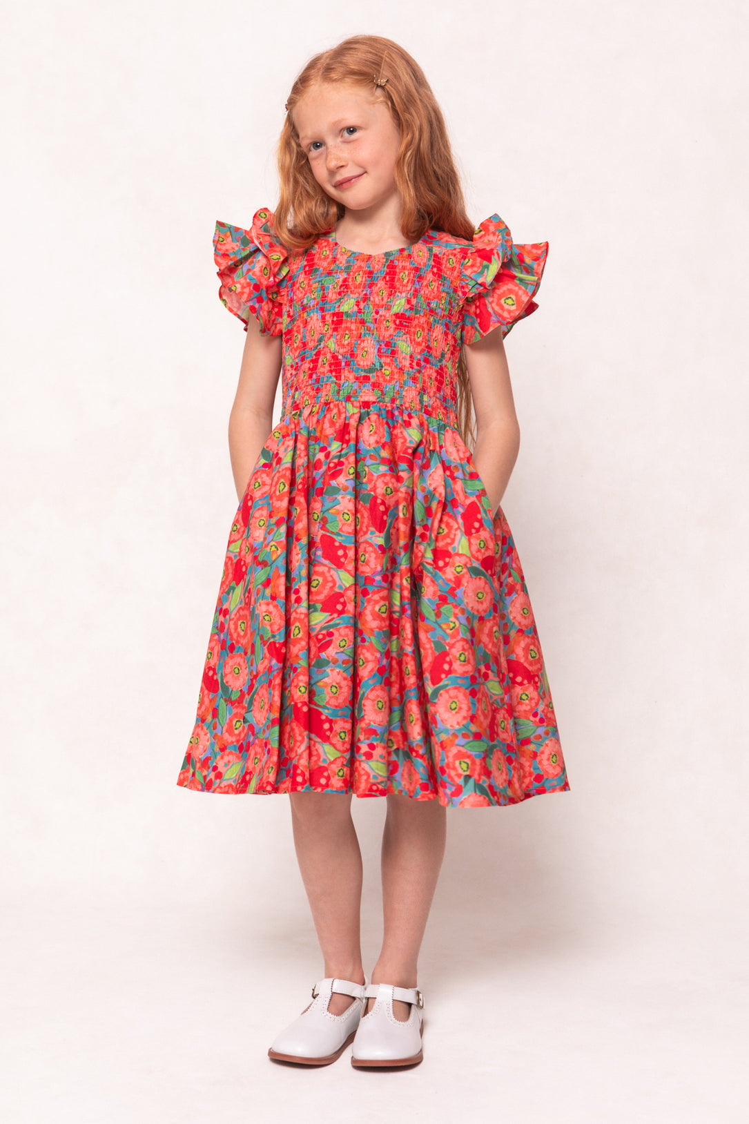 A young girl with long red hair wears the Ivy City Co Mini Hattie Dress in Poppy, featuring ruffled sleeves and pockets. She smiles in white shoes against a light background, hands tucked into the dress pockets.