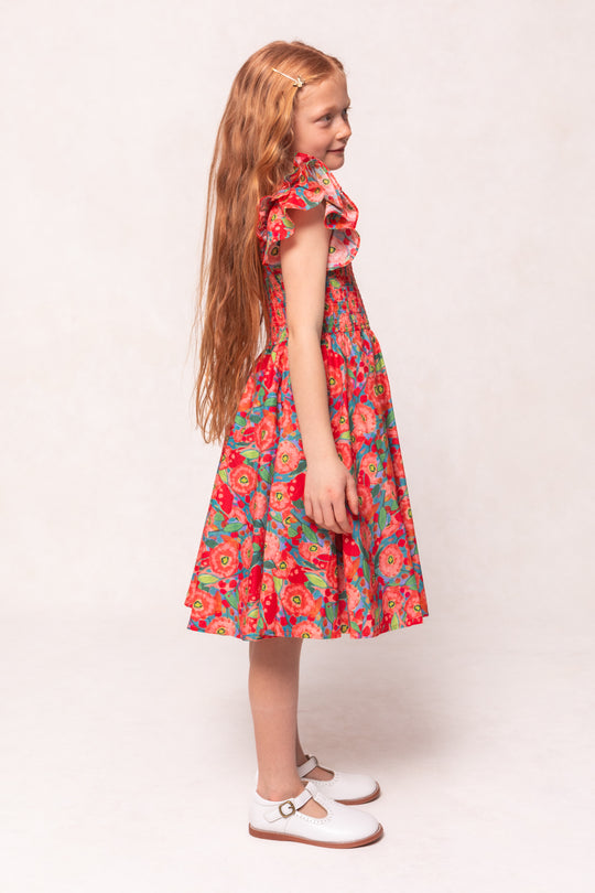 A young girl with long red hair stands in profile, wearing the Ivy City Co Mini Hattie Dress in Poppy with ruffled sleeves and white strap shoes, set against a plain light background.