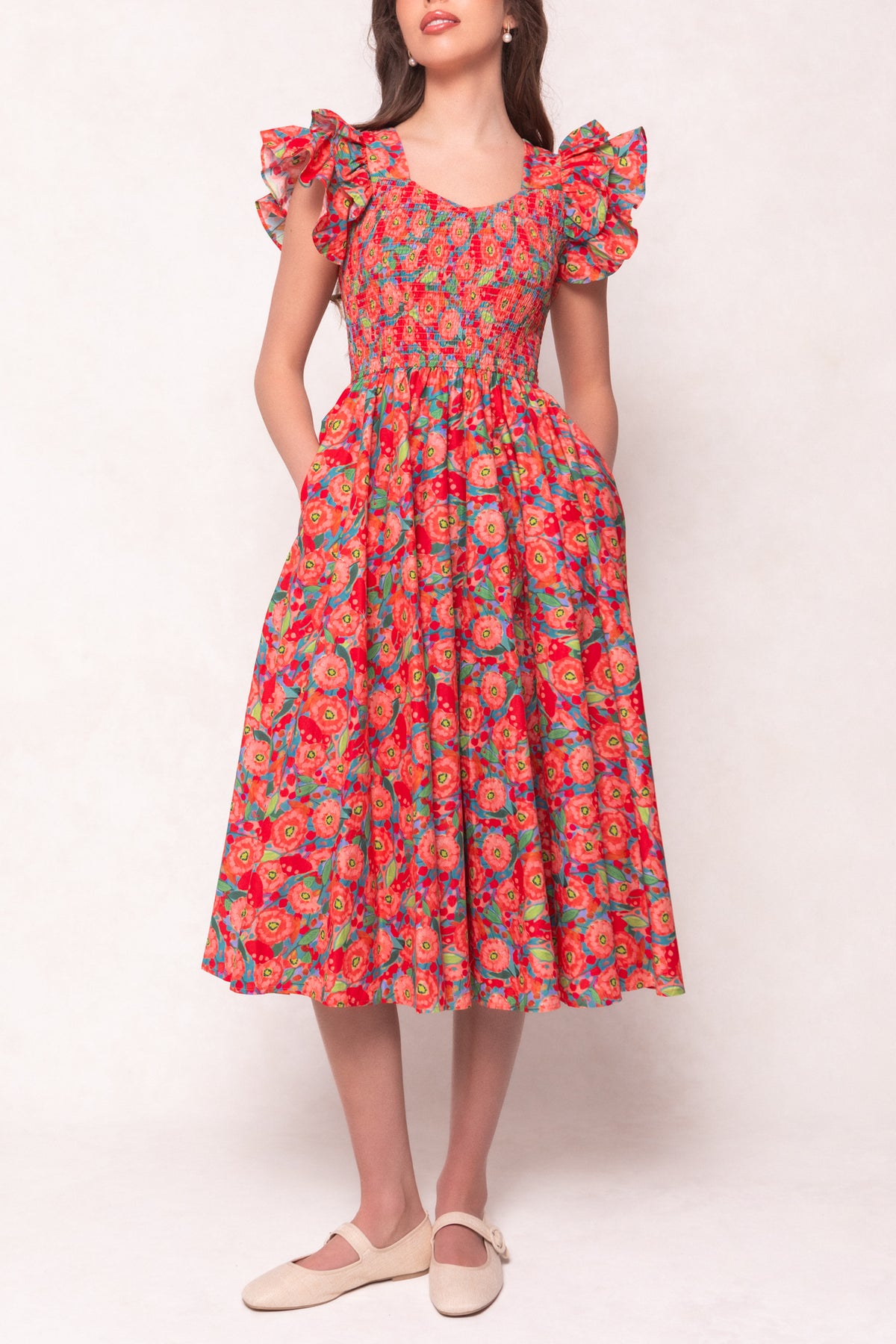 A woman wears the Ivy City Co Hattie Dress in Poppy, a cotton midi dress with ruffled cap sleeves and a smocked bodice. She pairs it with cream flats and stands against a light background.