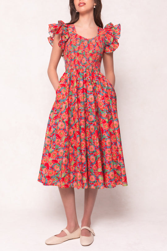 A woman wears the Ivy City Co Hattie Dress in Poppy, a cotton midi dress with ruffled cap sleeves and a smocked bodice. She pairs it with cream flats and stands against a light background.