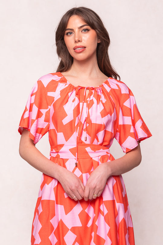 A woman with long brown hair wears the Ivy City Co Haven Dress in Geometric, a pink and orange cotton maxi dress featuring short flutter sleeves and a tie neckline, standing against a light background and smiling slightly at the camera.
