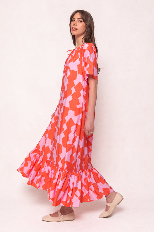 A woman poses sideways in the Ivy City Co Haven Dress in Geometric, featuring a pink and orange pattern, ruffled hem, short sleeves, and beige flats. The loose, ankle-length style is maternity friendly against a plain light background.
