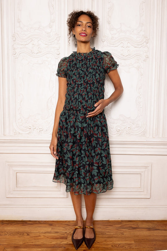 A woman wears the dark green Hayden Dress in Berries, a maternity-friendly midi featuring a smocked bodice, short puff sleeves, and a berry and leaf print.