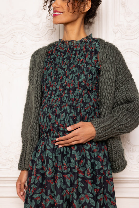 Woman wearing a patterned dress with a chunky knit cardigan against a textured white wall.