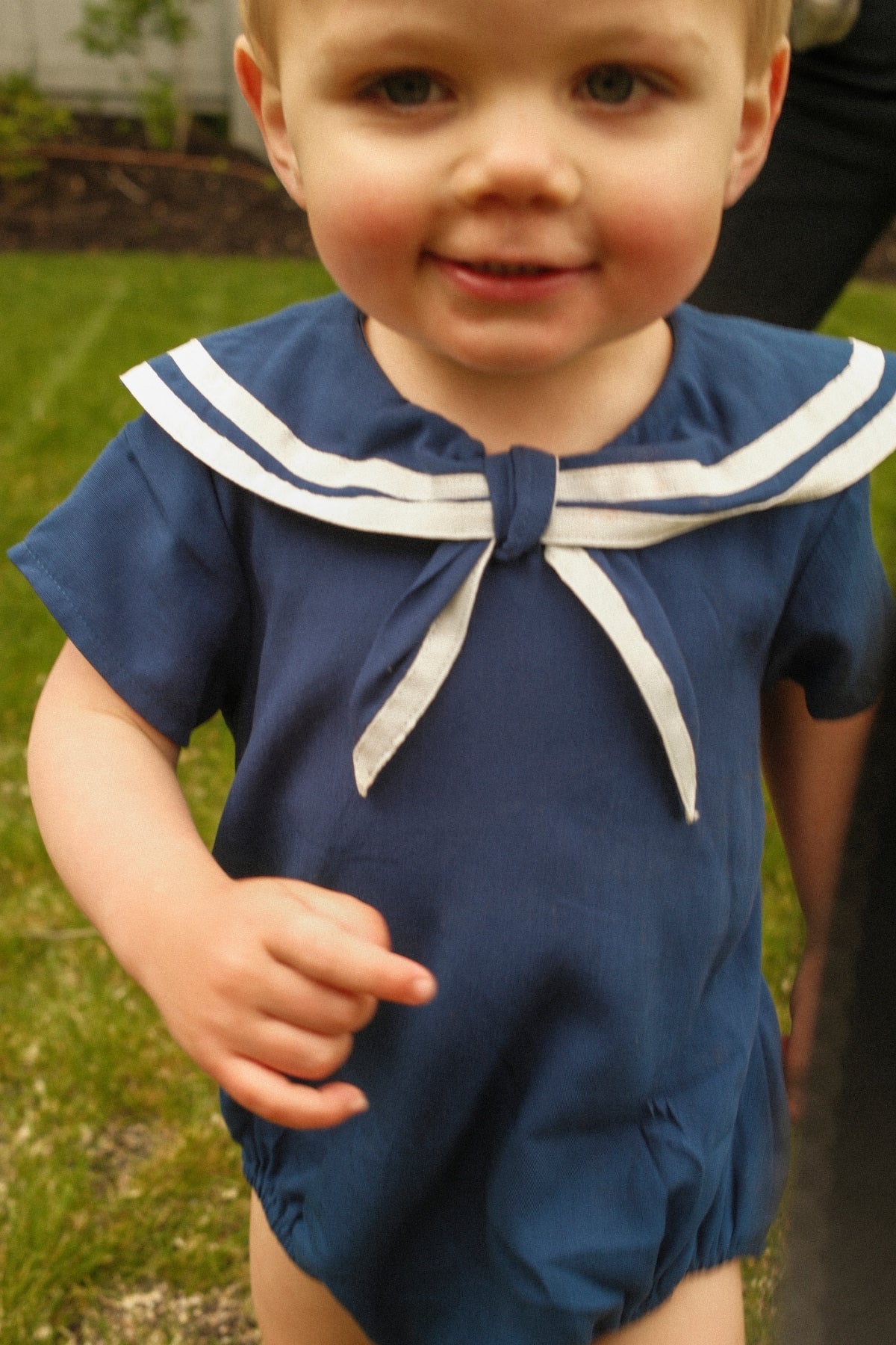 A toddler wearing the Ivy City Co Baby Sailor Romper in Navy with a crisp white sailor-style collar smiles while standing outside on the grass.