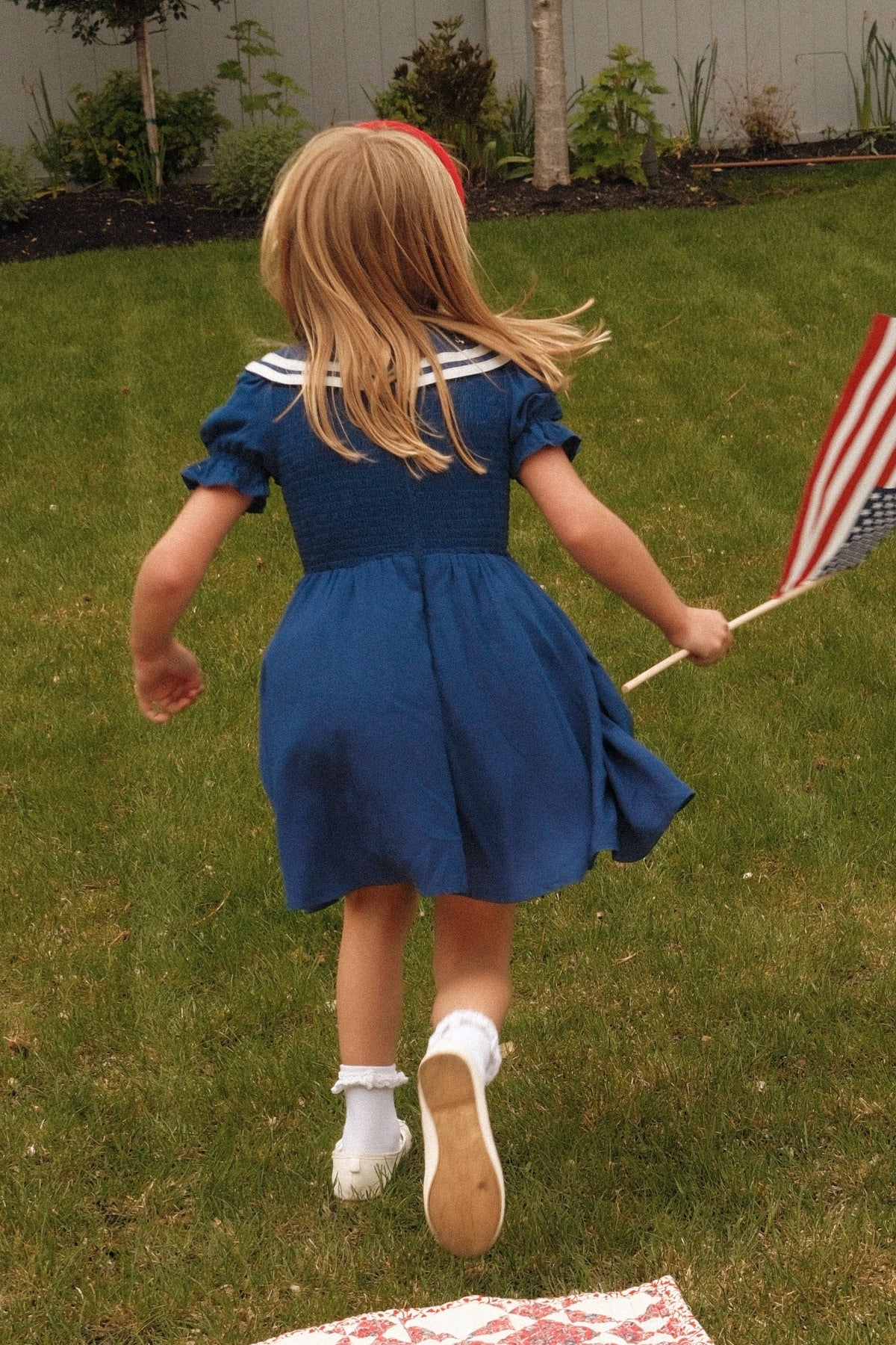 A young child wearing the Ivy City Co Mini Alice Dress in Navy runs on grass while holding an American flag, facing away from the camera.