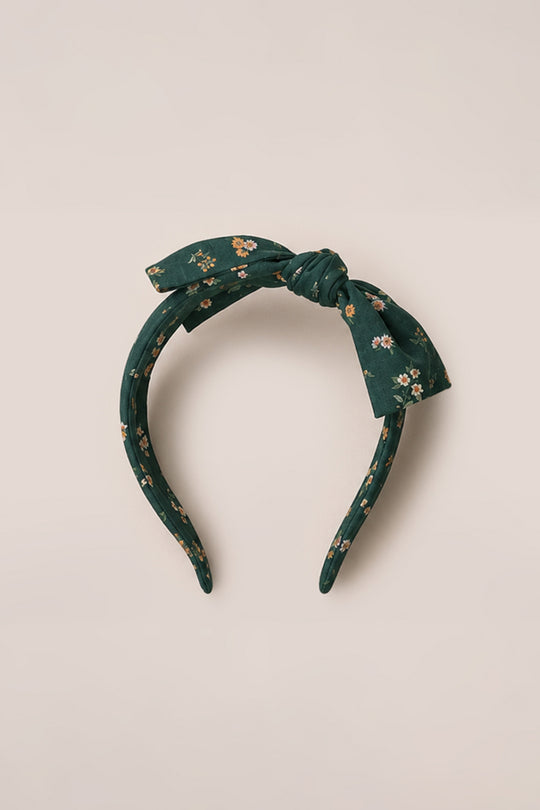 A dark green fabric headband from Ivy City Co, called Hair Bows in Evergreen Lovelorn Ditsy, features a tied bow at the top and is decorated with small yellow and pink floral prints in the signature Evergreen Lovelorn Ditsy pattern. The headband is displayed on a light beige background.