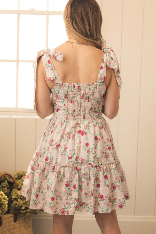 A woman stands indoors facing away, wearing the Ivy City Co Helene Dress in Vibrant Chintz—a light pink sundress featuring a floral chintz print, ruffled tiers, and tied shoulder straps. Sunlight filters through a window, and a basket of flowers rests on the floor nearby.