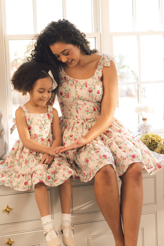 A woman and a young girl, both dressed in matching Ivy City Co Helene Dresses in Vibrant Chintz, sit together and smile as they look at something in the woman’s hand. Sunlight pours into the bright room from the large windows behind them.