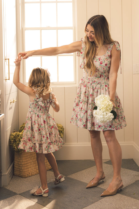 A woman and a young girl smile as they hold hands and dance in a sunlit room, both wearing matching Helene Dresses in Vibrant Chintz by Ivy City Co. The woman holds white flowers, while a basket of blooms rests by the window.