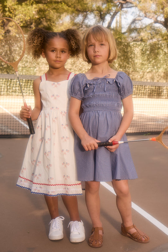 Two young girls stand on a tennis court holding rackets. One wears the Ivy City Co Mini Rae Dress in Blue Polka Dot with sandals, while the other sports a white cotton dress and sneakers. The net and trees are visible behind them.