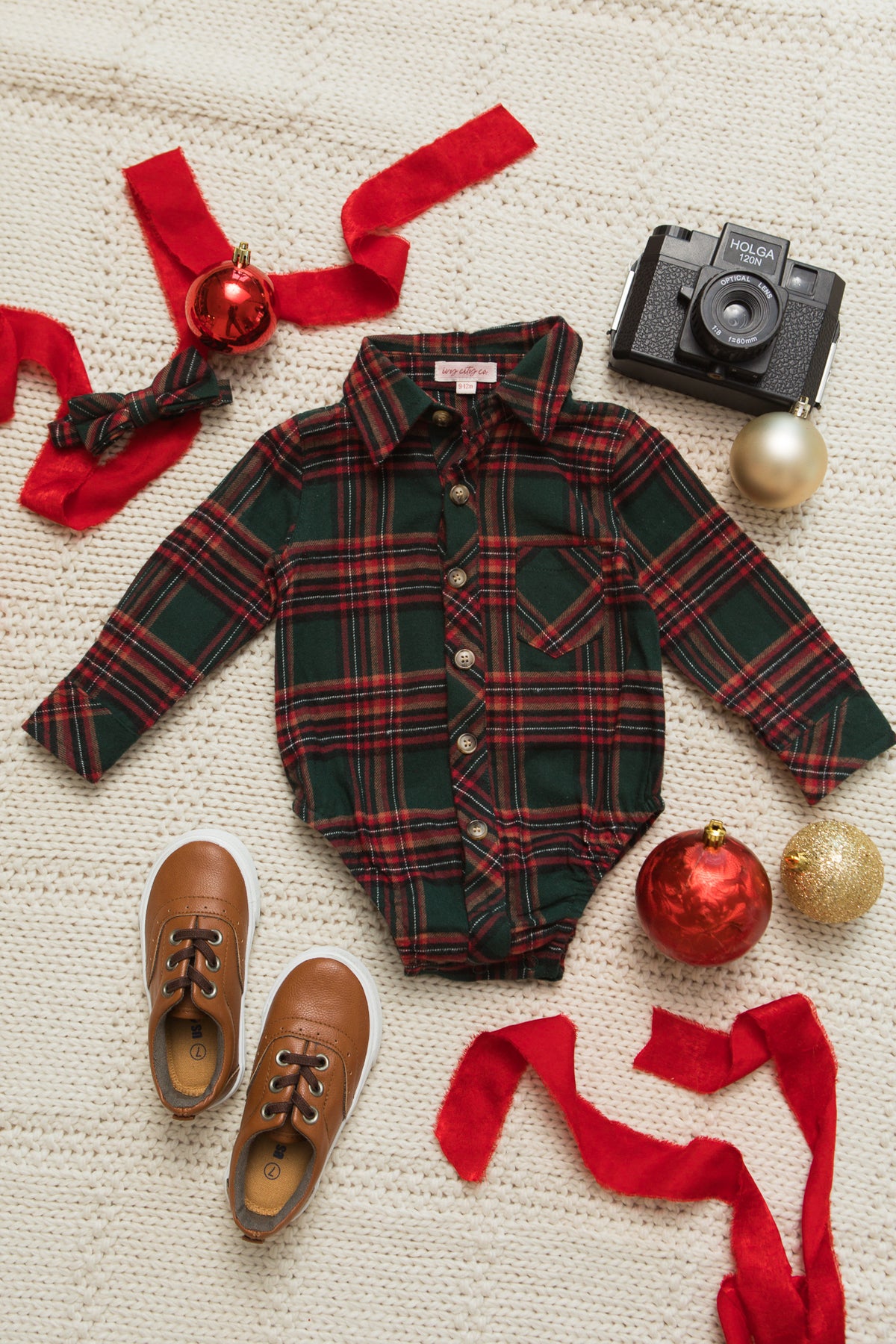The Ivy City Co Baby Boys John Romper in Evergreen Plaid, crafted from 100% cotton, is styled with brown baby shoes, a coordinating headband, festive ornaments, red ribbon, and a vintage camera—all beautifully arranged on a cream textured background for a charming holiday baby outfit.