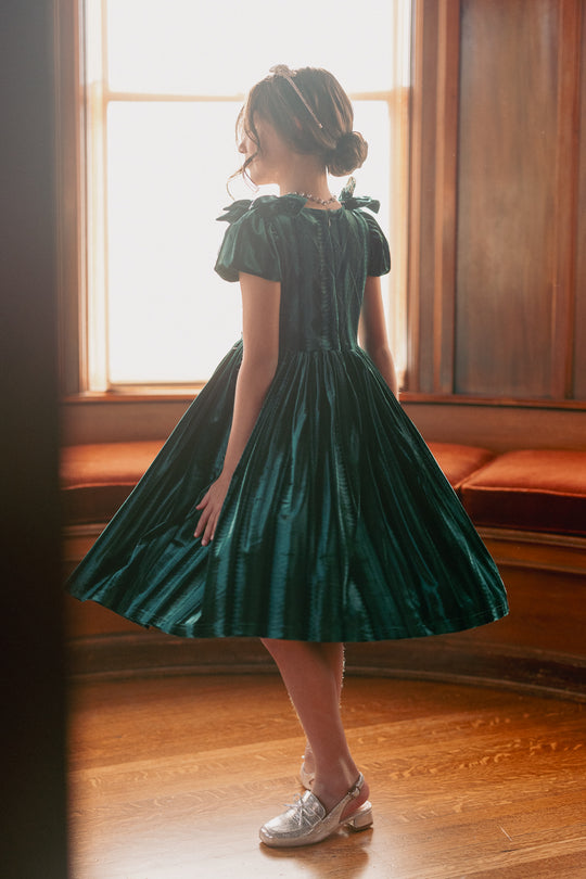 A girl wearing a green velvet mini dress with a square neckline, flutter sleeves, and diamond embroidery. She is standing indoors with a window behind her.