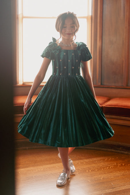 A girl wearing a green velvet mini dress with a square neckline, flutter sleeves, and diamond embroidery. She is standing indoors with a window behind her.