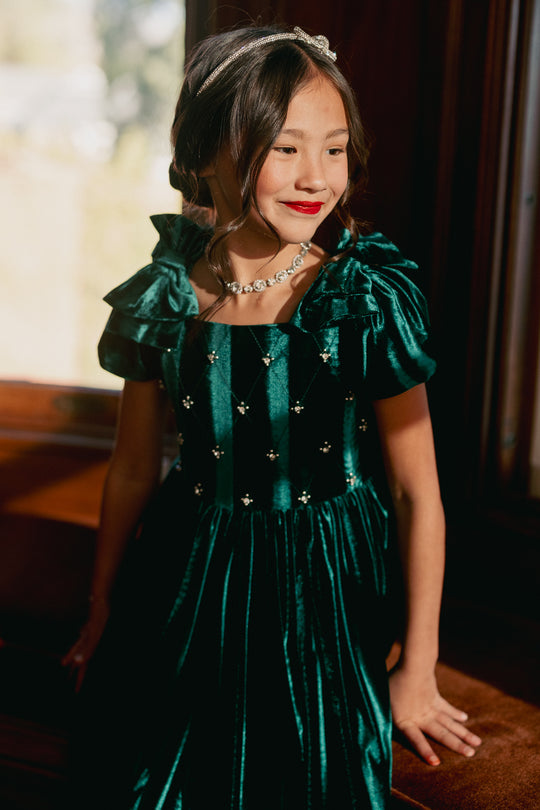 A girl wearing a green velvet mini dress with a square neckline, flutter sleeves, and diamond embroidery. She is sitting indoors with a window behind her.