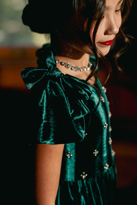 A girl wearing a green velvet mini dress with a square neckline, flutter sleeves, and diamond embroidery, with a velvet bo on the shoulder, standing indoors