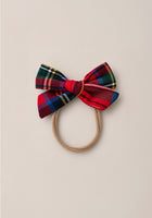 The Ivy City Co Hair Bows in Holiday Plaid features a tan elastic hair tie adorned with a large cotton flannel bow in a festive red, blue, yellow, green, and black plaid pattern, shown against a plain beige background.