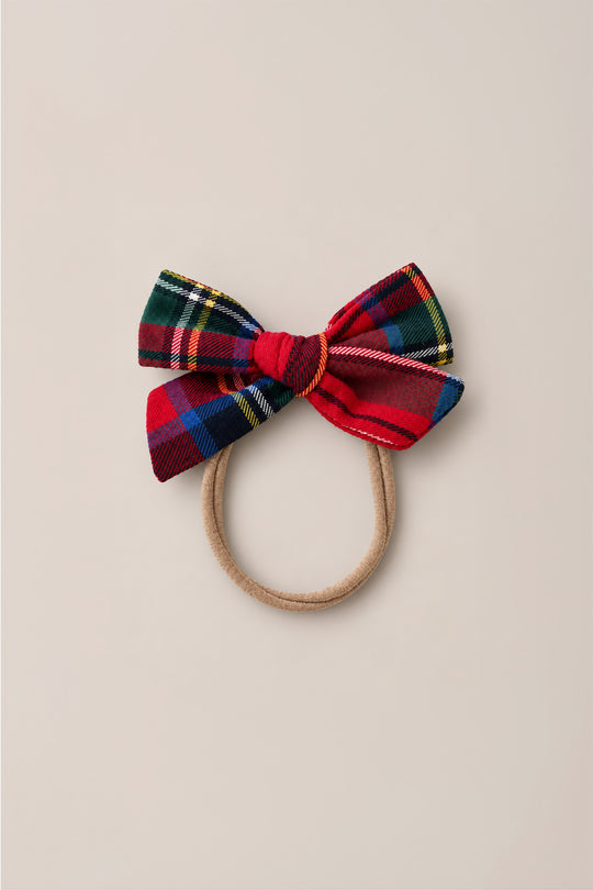 The Ivy City Co Hair Bows in Holiday Plaid features a tan elastic hair tie adorned with a large cotton flannel bow in a festive red, blue, yellow, green, and black plaid pattern, shown against a plain beige background.