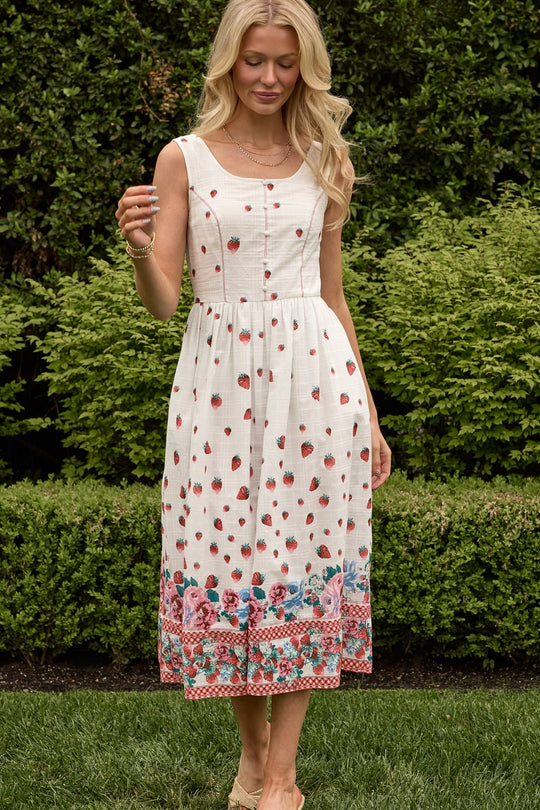 Wearing the Ivy City Co Sadie Dress in Strawberry Vintage Tier—a sleeveless cotton summer dress with red floral patterns and a colorful border—a woman stands on the grass among green bushes, looking down and smiling softly.