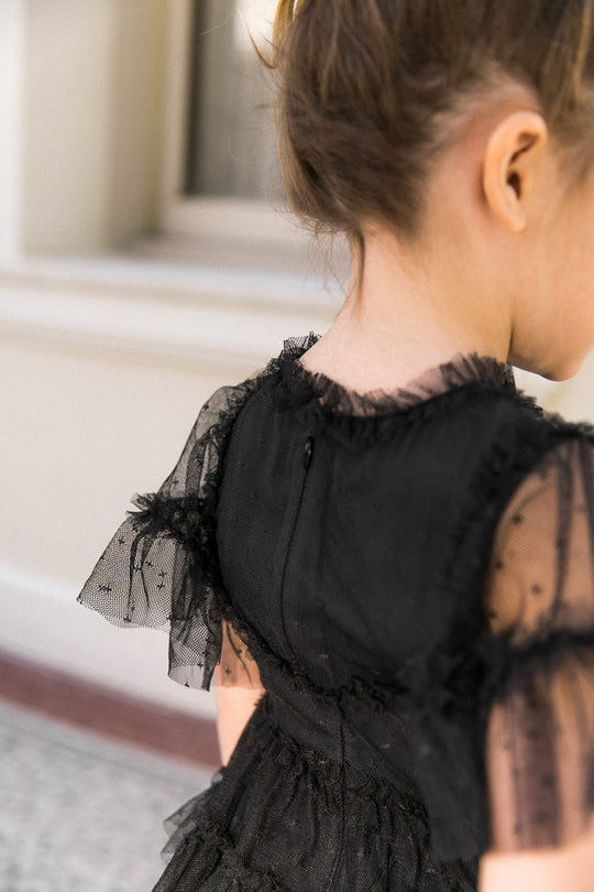 A young girl with light brown hair wears the Ivy City Co Mini Whimsical Dress in Black, featuring a sheer, ruffled bodice and polka-dot mesh flutter sleeves, standing outdoors with her back partially turned to the camera.