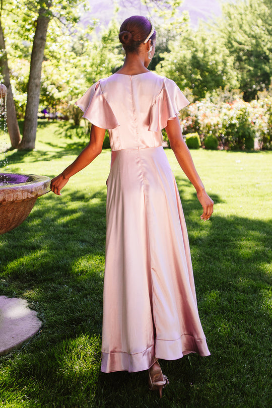 A woman wearing the Callie Dress in Blush Satin by Ivy City Co stands outdoors on green grass, her back to the camera. She is beside a fountain, surrounded by lush trees and sunlight.