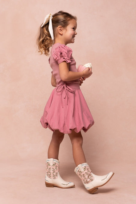 A young girl in a pink dress and Ivy City Co Mini Rosemary Cowgirl Boots with embroidered florals stands in profile, holding a doll against a soft pink background.