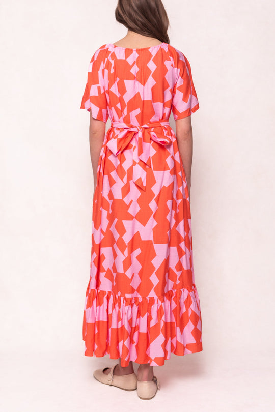 A person stands facing away, wearing Ivy City Co’s Haven Dress in Geometric, featuring a red and pink geometric pattern, ruffled hem, waist tie, and beige shoes.