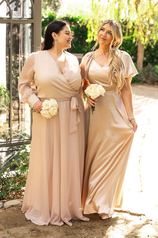 Wearing the Ivy City Co Tessie Dress in Champagne, two women stand outdoors holding white rose bouquets, smiling at each other with greenery and sunlight in the background.