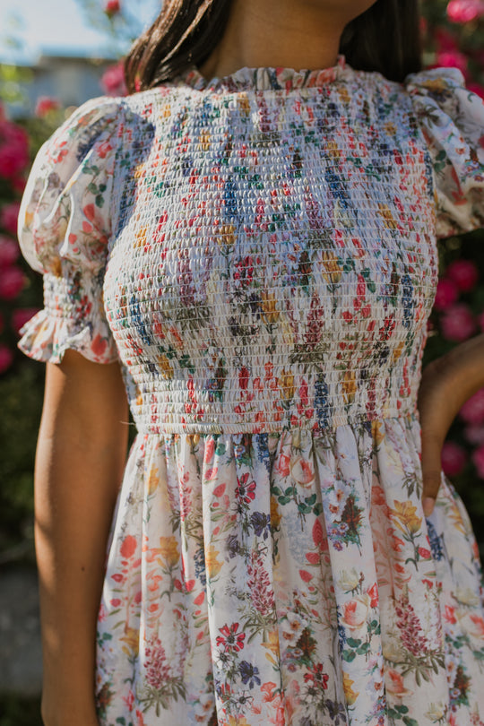 A woman wears the Ivy City Co Cordelia Floral Dress—light, floral, with puff sleeves and a smocked bodice—standing outdoors before pink flowers. The image is cropped from her shoulders to her knees.