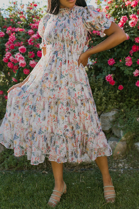 A woman in the Ivy City Co Cordelia Floral Dress, featuring short sleeves, ruffles, and a smocked bodice, stands in a rose garden holding one side of her dress. Her face is not visible.