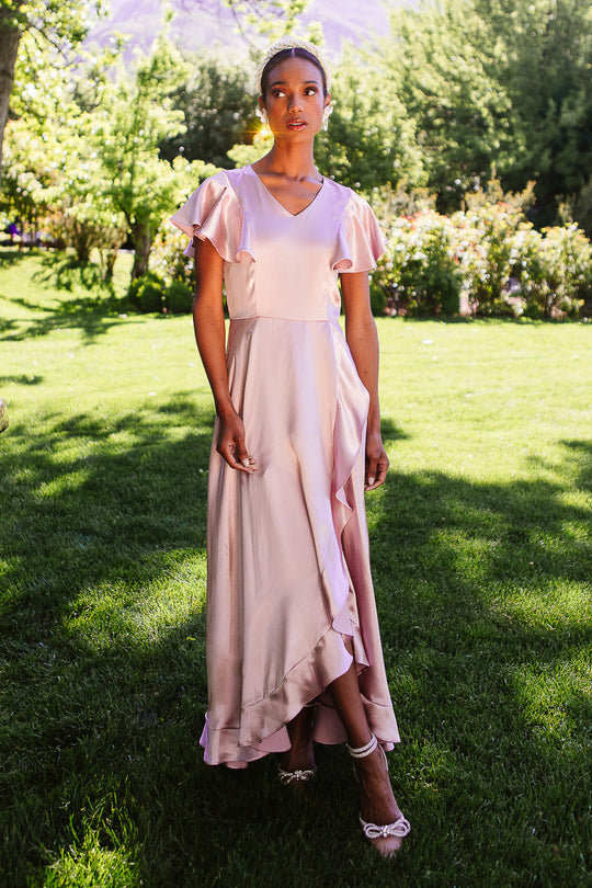A woman stands on the grass in sunlight, wearing the Ivy City Co Callie Dress in Blush Satin, featuring short sleeves and a ruffled wrap detail. She pairs it with ankle-strap heels, while green trees and bushes fill the background.