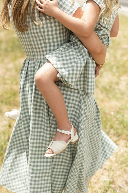 An adult and a young girl, both wearing the Ivy City Co Amber Dress in Sage Gingham, stand together on the grass outdoors. Their faces are not visible, and they both wear white sandals.