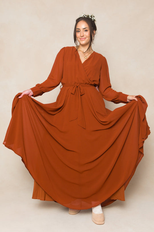 A woman wearing the Ivy City Co Andie Dress in Amber Chiffon gracefully holds out the sides of her flowing, long-sleeved maxi skirt. With a flower crown in her hair, she smiles while standing against a neutral background.