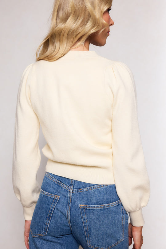 A woman with wavy blonde hair is seen from the back wearing the Illyria Sweater in Cream by Ivy City Co, featuring puffed shoulders, styled with blue high-waisted jeans for a relaxed look.