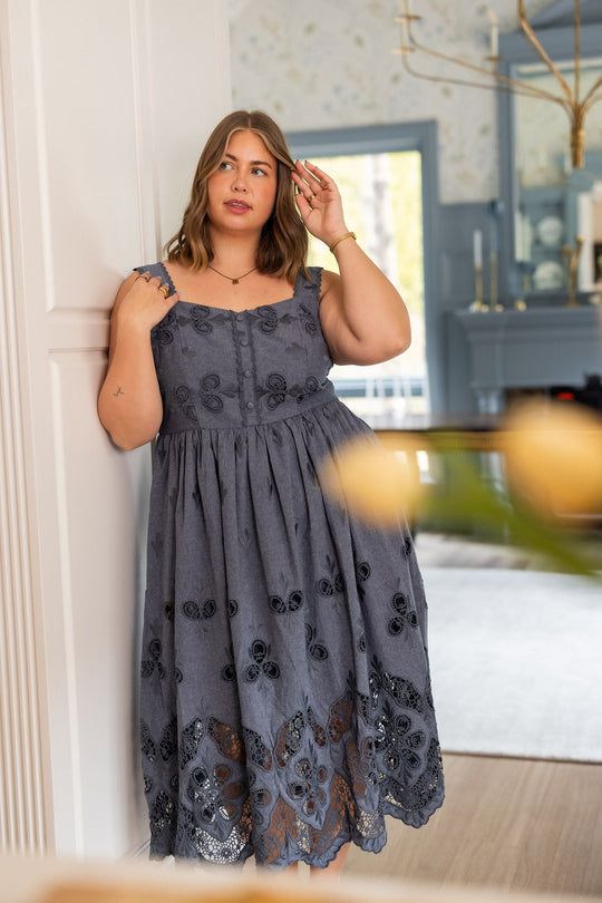 A woman in the Ivy City Co Penny Tank Midi Dress in Embroidered Chambray stands indoors by a white wall, touching her hair and gazing to the side. The sleeveless gray cotton dress features delicate eyelet embroidery, set against a stylish living room with light blue accents.