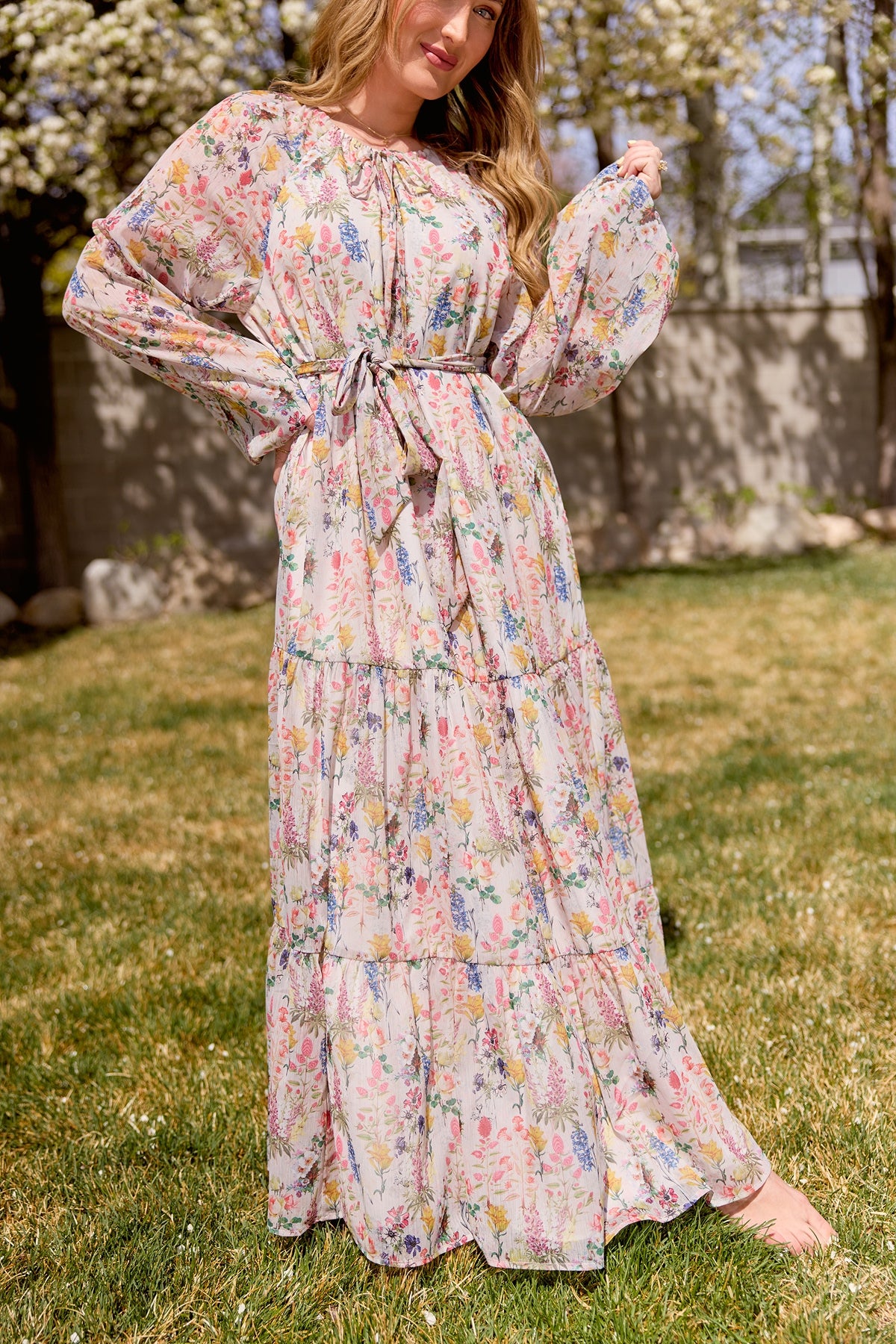 A woman stands barefoot on grass in the Ivy City Co Imogen Dress in Cordelia Floral—a flowy chiffon maxi with billowy sleeves and tied waist—surrounded by blooming trees for an effortlessly elegant, maternity-friendly look.