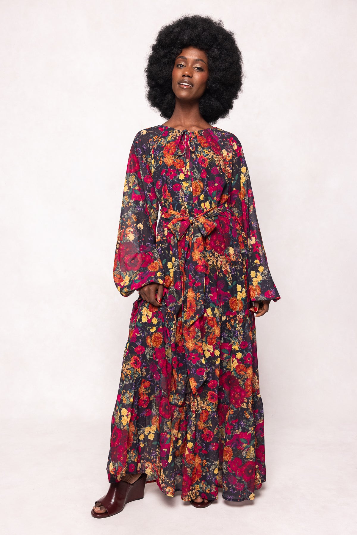 A woman with an afro hairstyle models the Ivy City Co Imogen Dress in Night Garden—a long sleeve floral dress with a vibrant red, yellow, and purple pattern, tied waist, and chic maternity-friendly style—against a light background.