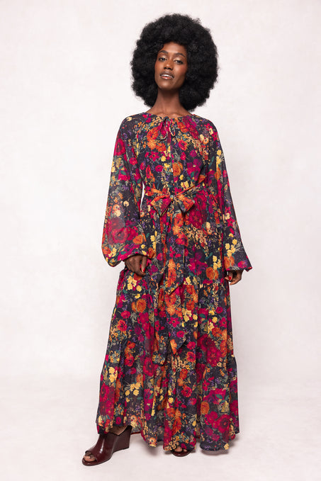 A woman with an afro hairstyle models the Ivy City Co Imogen Dress in Night Garden—a long sleeve floral dress with a vibrant red, yellow, and purple pattern, tied waist, and chic maternity-friendly style—against a light background.